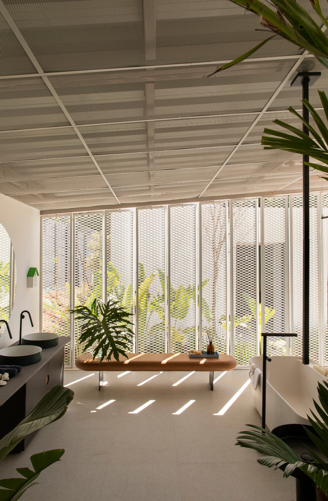 Banheiro com banheira solta, bancada de cubas escuras e luz filtrada pelo metal expandido
