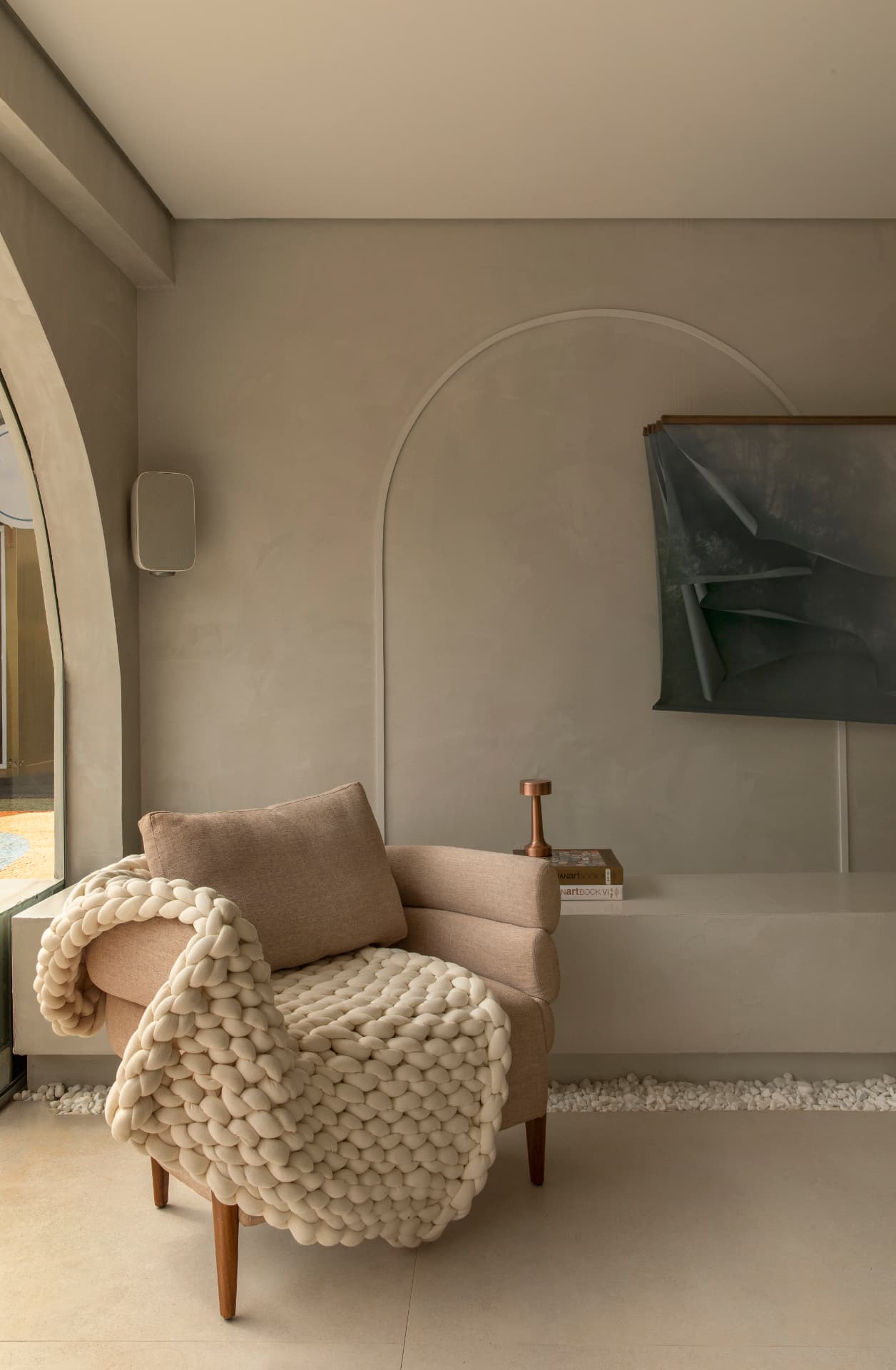Poltrona bege com manta trançada, nicho em arco e obra de arte