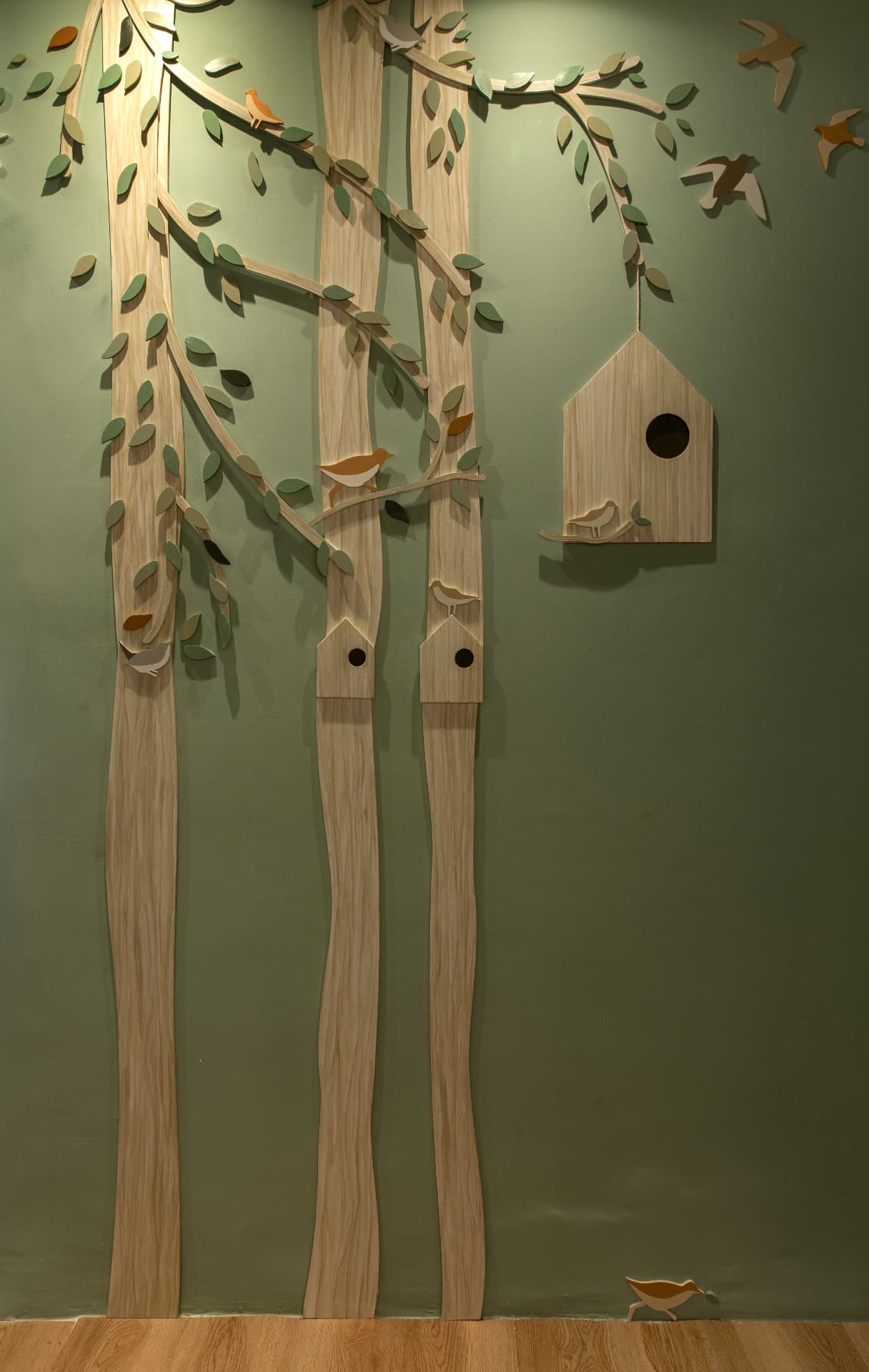 Painel de marcenaria com troncos, folhas e casinha de passarinho em parede verde